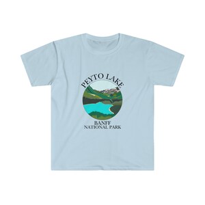 May include: Light blue t-shirt featuring a circular graphic of Peyto Lake in Banff National Park. The graphic depicts a turquoise lake surrounded by green mountains and a snowy peak.
