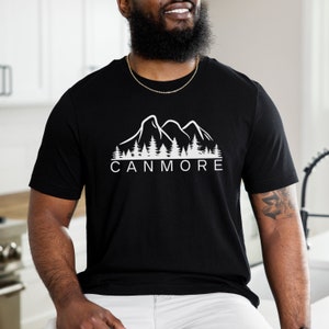 May include: Black t-shirt featuring a white graphic of mountains and trees. The text "CANMORE" is printed below the graphic.