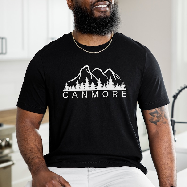 May include: Black t-shirt featuring a white graphic of mountains and trees. The text "CANMORE" is printed below the graphic.