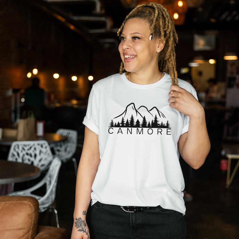 May include: White t-shirt with a black mountain range and forest graphic and the text "Canmore" printed on the front.