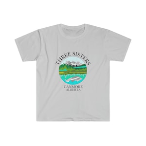 May include: A light grey t-shirt featuring a circular graphic of the Three Sisters mountains in Canmore, Alberta. The graphic is in shades of green, blue, and grey, with the text "Three Sisters" and "Canmore Alberta" in black.