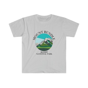 May include: A light grey t-shirt featuring a circular graphic of a mountain range in green, blue and grey colours. The graphic is surrounded by the text "MOUNT RUNDLE" in black. Below the graphic is the text "BANFF NATIONAL PARK" in black.