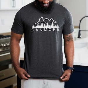 May include: A dark heather grey t-shirt with a white graphic of mountains and trees. The text "CANMORE" is printed below the graphic.