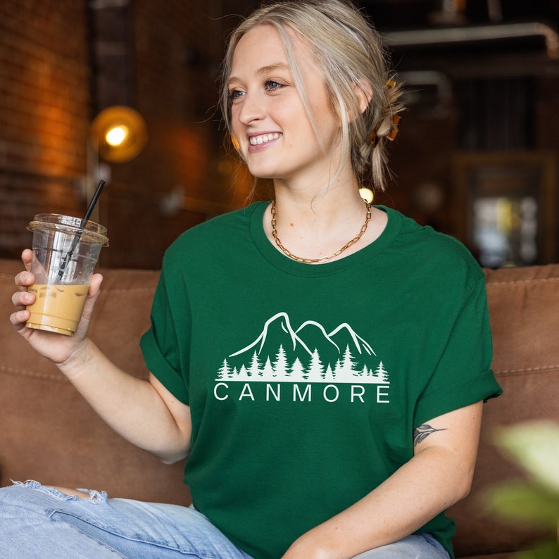May include: A green t-shirt featuring a white graphic of mountains and trees. The text "Canmore" is printed below the graphic.