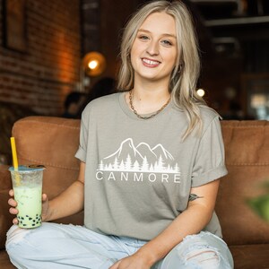 May include: A woman wearing a light grey t-shirt with a white mountain and trees graphic and the text "Canmore". She is holding a green bubble tea in her left hand.