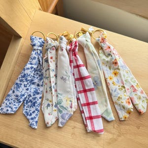 May include: A collection of six fabric keychains with gold-colored rings. The fabric patterns include blue floral, white floral, and red plaid designs. The keychains are arranged on a wooden surface.