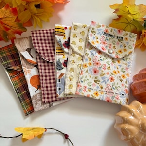 May include: A collection of fabric pouches with various patterns, including plaid, pumpkins, chickens, and floral designs. One pouch features the text "JEREMIAH 29:11" and "PEACE FILLED LIFE WITH JESUS". The pouches are arranged with autumn leaves and decorative pumpkins.