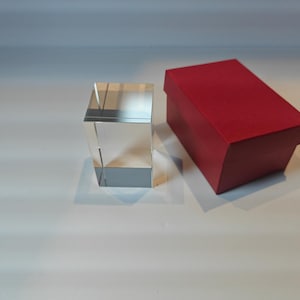 Op de afbeelding: Een helder, rechthoekig kristallen blok naast een rode rechthoekige doos. Het kristallen blok is transparant en reflecteert licht. De rode doos heeft een bijpassend rood deksel.