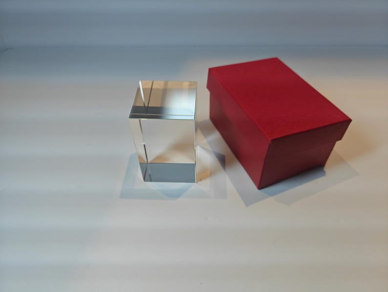 Op de afbeelding: Een helder, rechthoekig kristallen blok naast een rode rechthoekige doos. Het kristallen blok is transparant en reflecteert licht. De rode doos heeft een bijpassend rood deksel.