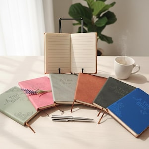 Könnte beinhalten: Ein offenes Notizbuch auf einem schwarzen Ständer, umgeben von sechs geschlossenen Notizbüchern in verschiedenen Farben: Salbeigrün, Rosa, Grau, Orange, Dunkelgrün und Blau. Ein silberner Stift liegt daneben. Eine weiße Tasse, aus der Dampf aufsteigt.