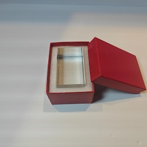 Op de afbeelding: Een helder, rechthoekig glazen blok wordt gepresenteerd in een open rode geschenkdoos met een bijpassend rood deksel. Het blok is gecentreerd op een witte schuimrubberen inzet. De doos staat op een wit oppervlak.