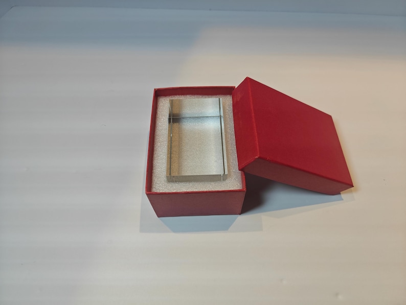 Op de afbeelding: Een helder, rechthoekig glazen blok wordt gepresenteerd in een open rode geschenkdoos met een bijpassend rood deksel. Het blok is gecentreerd op een witte schuimrubberen inzet. De doos staat op een wit oppervlak.