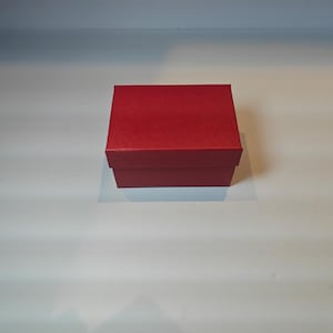 Op de afbeelding: Een kleine, vierkante, rode geschenkdoos met een bijpassende deksel. De doos staat op een wit oppervlak. De doos is gemaakt van een textuur.
