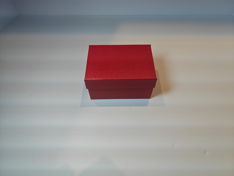 Op de afbeelding: Een kleine, vierkante, rode geschenkdoos met een bijpassende deksel. De doos staat op een wit oppervlak. De doos is gemaakt van een textuur.