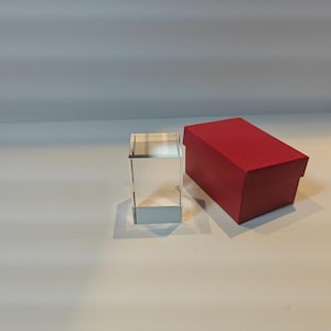 Op de afbeelding: Een helder, rechthoekig glazen displayblok staat naast een rode rechthoekige doos. Het displayblok is transparant. De rode doos heeft een bijpassend deksel.