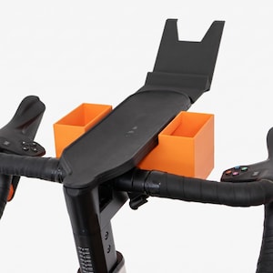 May include: A black bicycle handlebar assembly featuring a central black tray with an upright back support and two bright orange rectangular storage bins attached to the sides. The handlebars are wrapped in black tape with integrated control buttons.