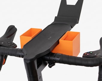 May include: A black bicycle handlebar assembly featuring a central black tray with an upright back support and two bright orange rectangular storage bins attached to the sides. The handlebars are wrapped in black tape with integrated control buttons.