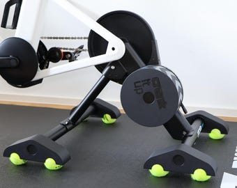 Pode incluir: Uma estrutura de bicicleta estacionária branca fixada a um rolo de treino preto. A base do rolo possui quatro pés de suporte pretos, cada um estabilizado por uma bola de ténis amarela brilhante. Uma tampa cinzenta circular exibe o texto Crank It Up.