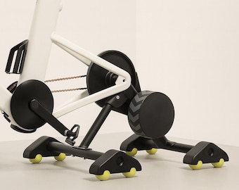 May include: A white indoor stationary exercise bike mounted on a black trainer stand. The bike features a black seat, black pedals, and a black flywheel. The trainer stand is supported by black legs with small, bright yellow wheels at the base.