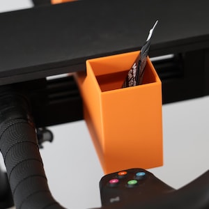 May include: A bright orange rectangular storage bin attached to a black exercise bike handlebar assembly. The bin features a smooth, matte finish and holds a small packet. The handlebar has a textured black grip and a control panel with buttons.