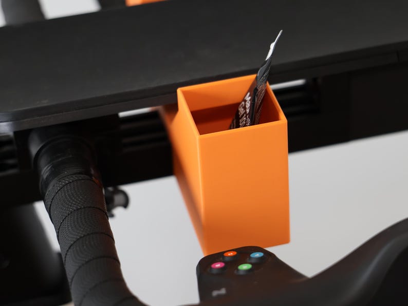 May include: A bright orange rectangular storage bin attached to a black exercise bike handlebar assembly. The bin features a smooth, matte finish and holds a small packet. The handlebar has a textured black grip and a control panel with buttons.