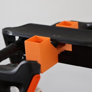 May include: A black and orange accessory mount attached to a handlebar assembly. The mount features two bright orange square-shaped holders and a flat black platform, integrated with black textured grips and mechanical components on the handlebars.