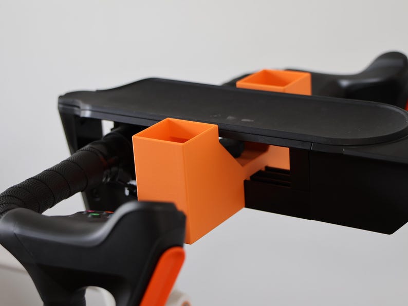 May include: A black and orange accessory mount attached to a handlebar assembly. The mount features two bright orange square-shaped holders and a flat black platform, integrated with black textured grips and mechanical components on the handlebars.