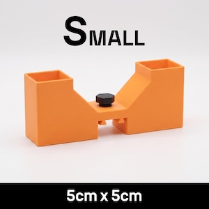 May include: An orange plastic tool with two square openings and a central black knob. The text SMALL is printed above the tool, and the text 5cm x 5cm is printed below. The tool features a bridge-like structure with two vertical columns.