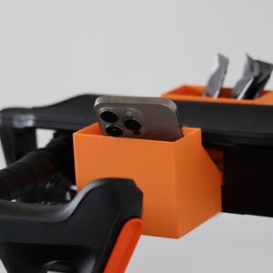 May include: A bright orange plastic storage bin attached to a black exercise bike handlebar assembly. The bin holds a gray smartphone in an upright position, while a second smaller orange bin behind it contains two dark rectangular packets.