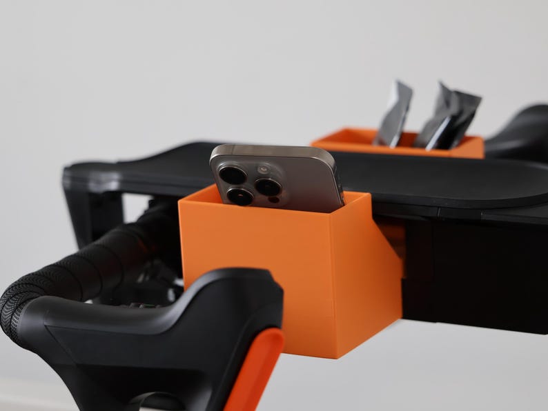 May include: A bright orange plastic storage bin attached to a black exercise bike handlebar assembly. The bin holds a gray smartphone in an upright position, while a second smaller orange bin behind it contains two dark rectangular packets.