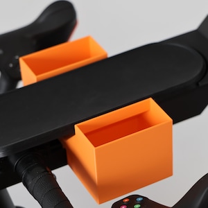 May include: A black exercise bike accessory tray featuring two bright orange rectangular storage compartments attached to a flat, elongated black surface that mounts directly onto the handlebars of a stationary cycling machine.