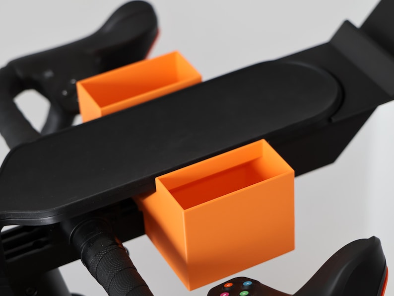 May include: A black exercise bike accessory tray featuring two bright orange rectangular storage compartments attached to a flat, elongated black surface that mounts directly onto the handlebars of a stationary cycling machine.