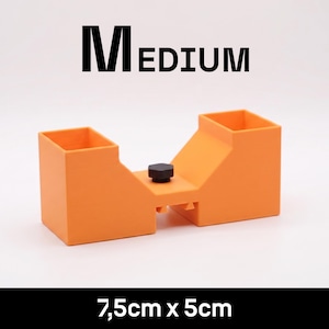 May include: An orange plastic tool with two square openings and a central black knob, labeled MEDIUM and 7.5cm x 5cm. The object features a symmetrical design with a recessed center section between the two vertical square-shaped compartments.