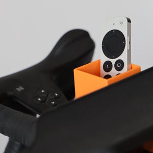 May include: A bright orange rectangular holder attached to black exercise equipment, containing a silver and black remote control with a circular navigation pad, power button, and icons for back and home functions on its front surface.