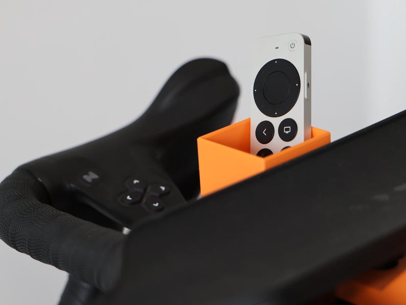 May include: A bright orange rectangular holder attached to black exercise equipment, containing a silver and black remote control with a circular navigation pad, power button, and icons for back and home functions on its front surface.