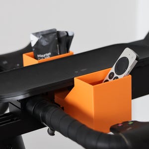 May include: Two bright orange plastic storage containers attached to the black handlebars of an exercise bike. One container holds a silver and black remote control, while the other holds several black Maurten Gel 100 energy gel packets.