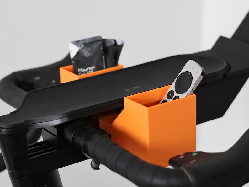 May include: Two bright orange plastic storage containers attached to the black handlebars of an exercise bike. One container holds a silver and black remote control, while the other holds several black Maurten Gel 100 energy gel packets.