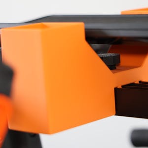 May include: A bright orange plastic mounting bracket with a rectangular open-top compartment, featuring a black textured adjustment knob positioned underneath a flat black surface, designed for secure attachment to a larger mechanical structure.