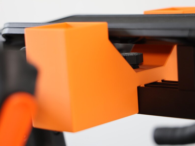 May include: A bright orange plastic mounting bracket with a rectangular open-top compartment, featuring a black textured adjustment knob positioned underneath a flat black surface, designed for secure attachment to a larger mechanical structure.