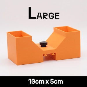 May include: An orange plastic tool with two rectangular openings and a central black knob. The text LARGE is printed above the object, and the text 10cm x 5cm is printed below. The object features a bridge-like structure with a hollow center.