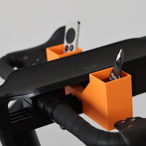 May include: Two bright orange rectangular storage holders attached to the black handlebars of an exercise bike. One holder contains a white remote control, and the other holds a small black and white packet labeled Maurten Gel 100.