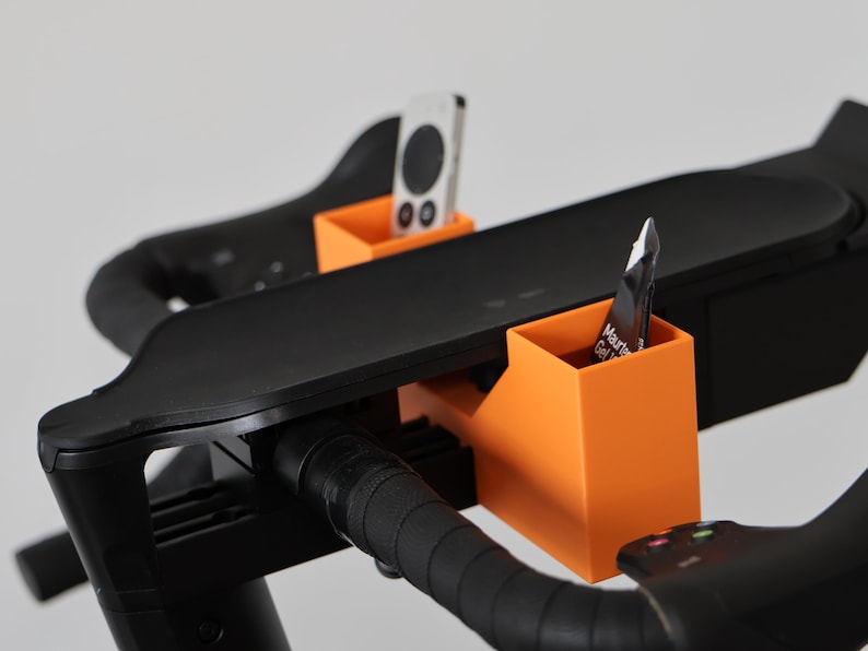 May include: Two bright orange rectangular storage holders attached to the black handlebars of an exercise bike. One holder contains a white remote control, and the other holds a small black and white packet labeled Maurten Gel 100.