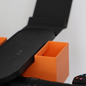 May include: A black plastic accessory mount featuring two rectangular orange storage bins attached to the sides. The central black component has a flat, elongated surface with a vertical back panel, designed to hold items securely on a handlebar.