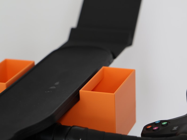 May include: A black plastic accessory mount featuring two rectangular orange storage bins attached to the sides. The central black component has a flat, elongated surface with a vertical back panel, designed to hold items securely on a handlebar.