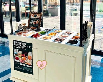 May include: A white wooden cart with a variety of toppings for charcuterie, including olives, cheese, and crackers. The cart has a sign that says "Elegance on Wheels & Flavor in Every Bite." and a pink heart with the text "slc" on it.