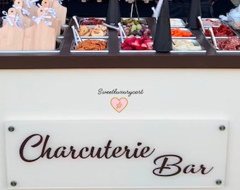 May include: A white cart with a sign that says "Charcuterie Bar" in black cursive lettering. The cart is filled with various food items, including fruit, cheese, crackers, and meat. The cart is decorated with a pink heart with the text "Sweetluxurycart" in black.