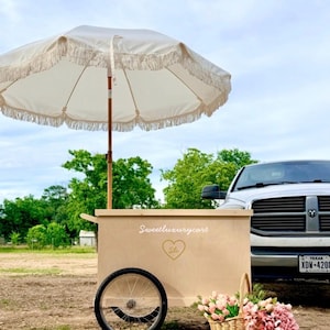 May include: A beige wooden cart with a cream-colored fringed umbrella. The cart has a single wheel and a basket of pink flowers. The text "Sweetluxurycart" and a heart logo are visible on the cart.