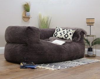 May include: A large, brown corduroy bean bag chair with a black and white patterned pillow and a book on top. The chair is sitting on a wooden floor with a black and white patterned rug in front of it.