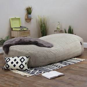 May include: A large, beige corduroy bean bag chair with a grey faux fur throw on top. The bean bag is sitting on a black and white patterned rug on a wooden floor.