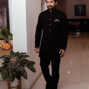 May include: A man in a black suit with a mandarin collar, black pants, and black shoes. The suit has a pocket square with a colorful pattern. The man is walking in a well-lit hallway.